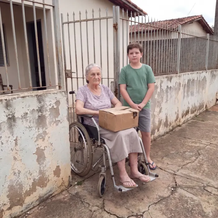 Neusa na cadeira de rodas em frente ao portão de casa, com a caixa de doces no colo, ao lado do neto Mateus